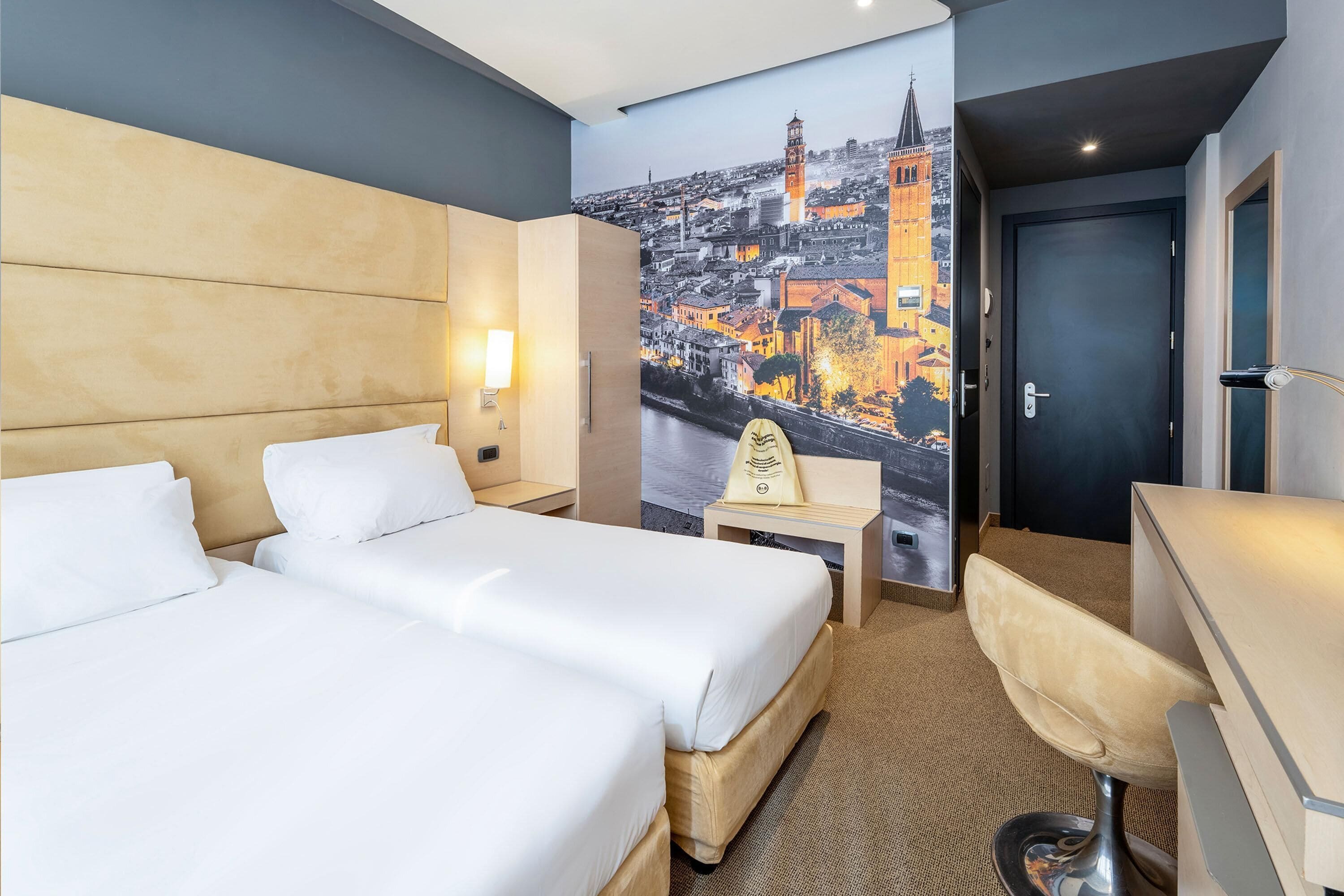 A hotel room with two beds, a desk, a chair, a nightstand, and a wall mural of a cityscape.