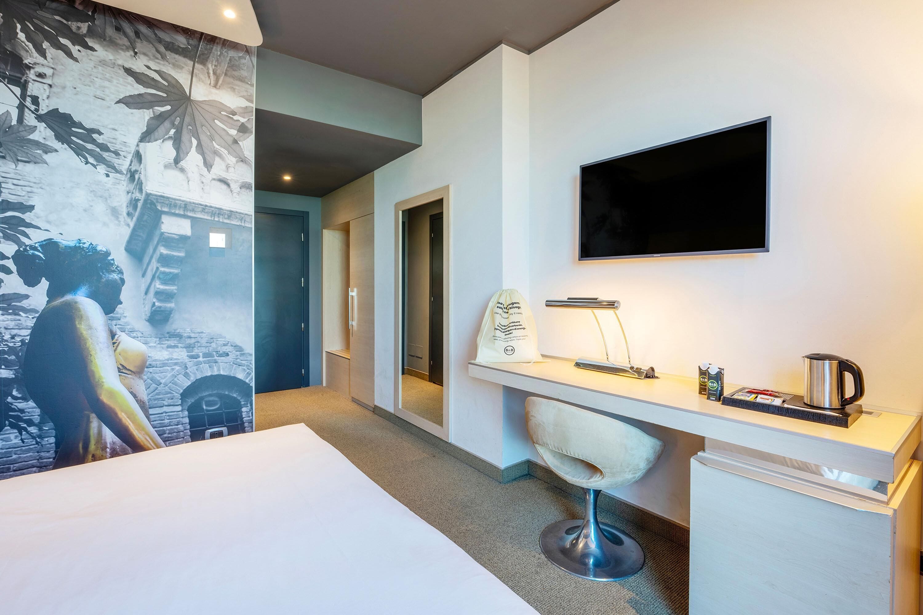 Ein Hotelzimmer mit einem Bett, einem Schreibtisch mit Stuhl, einem Fernseher und einer Wand mit Wandgemälde.