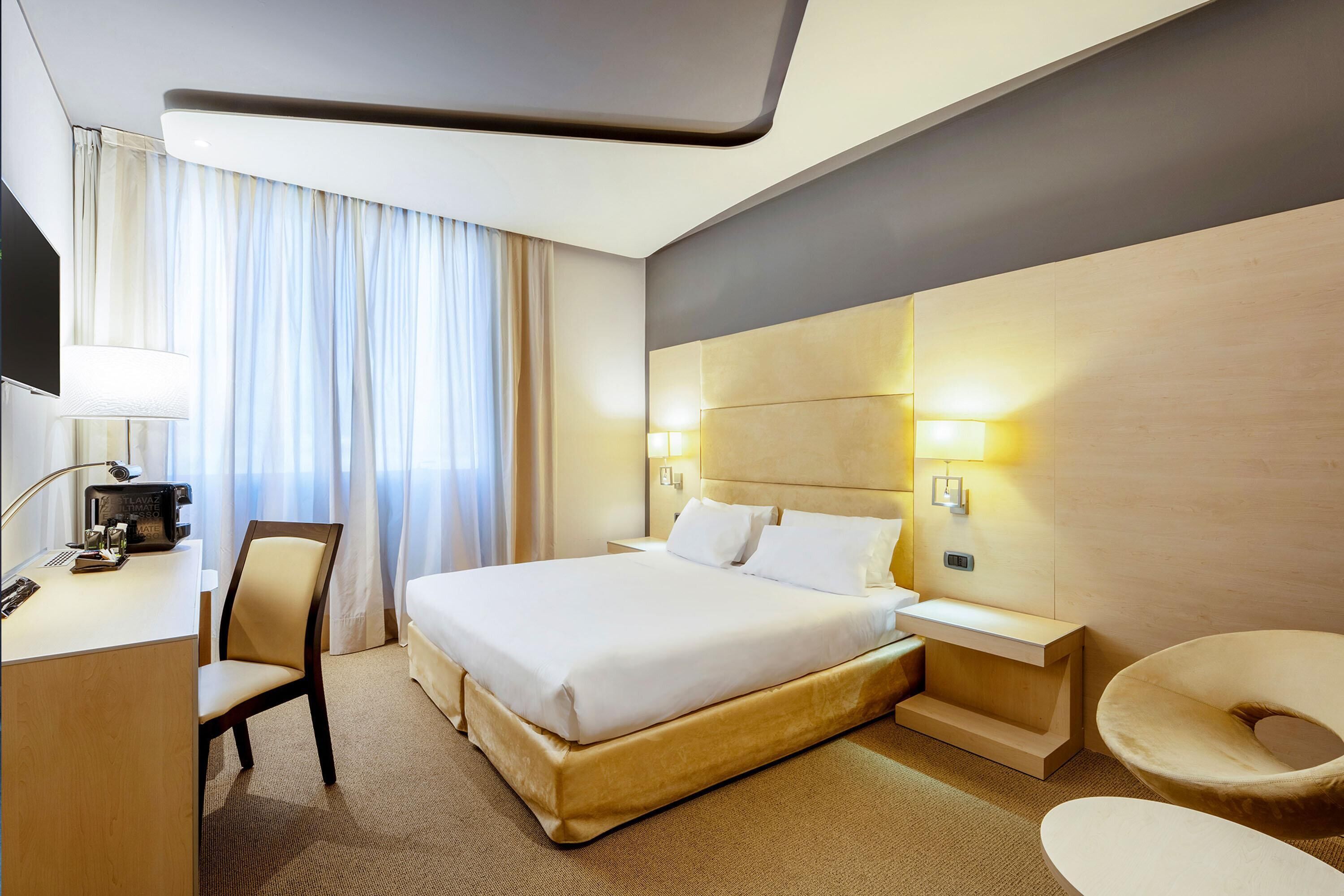 Habitación de hotel con cama, escritorio con lámpara, silla, mesita de noche con lámpara y un ventanal con cortinas.
