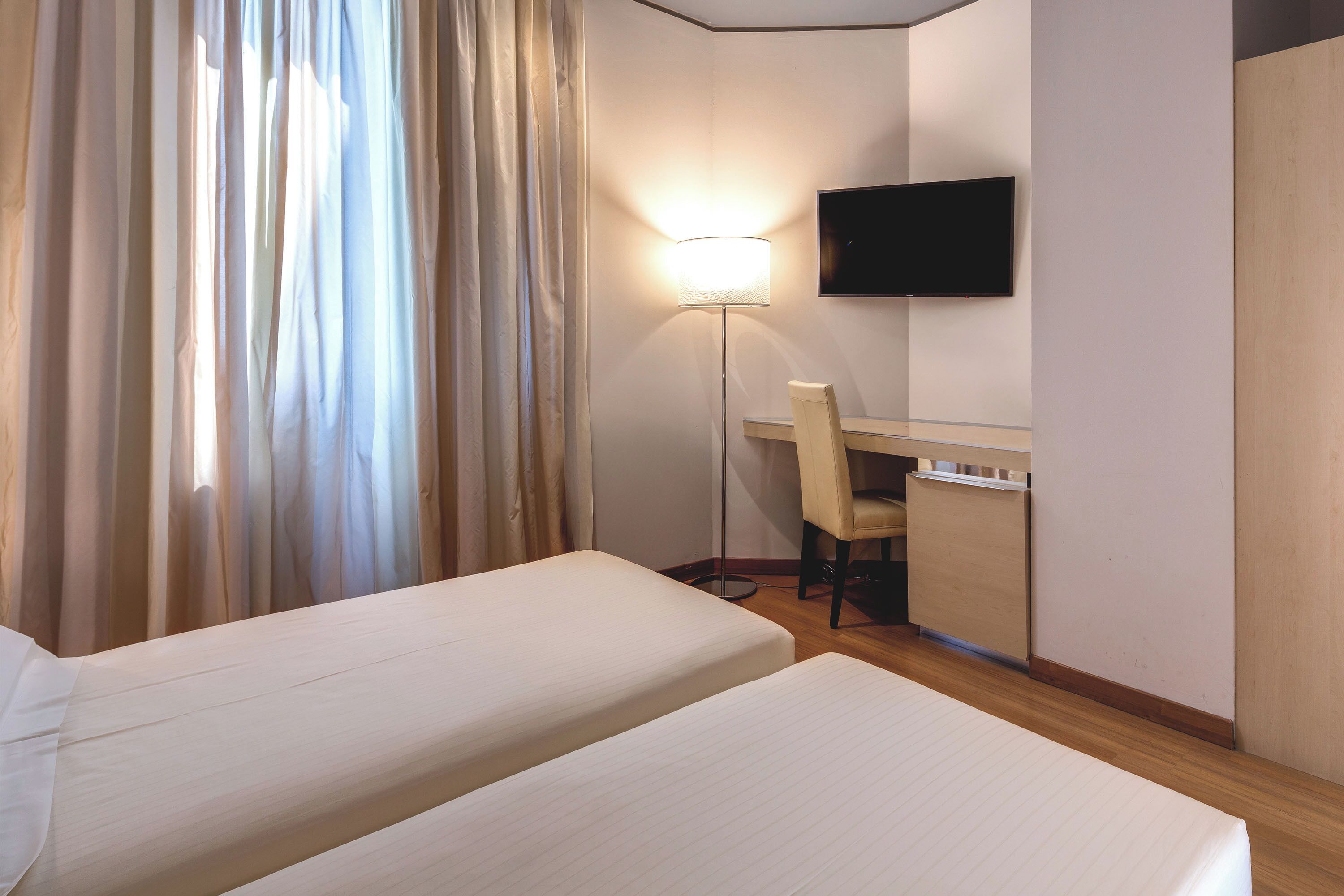 Hotellihuone, jossa on kaksi sänkyä, työpöytä, tuoli, televisio ja lattiavalaisin.