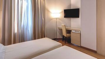 Hotellihuone, jossa on kaksi sänkyä, työpöytä, tuoli, televisio ja lattiavalaisin.