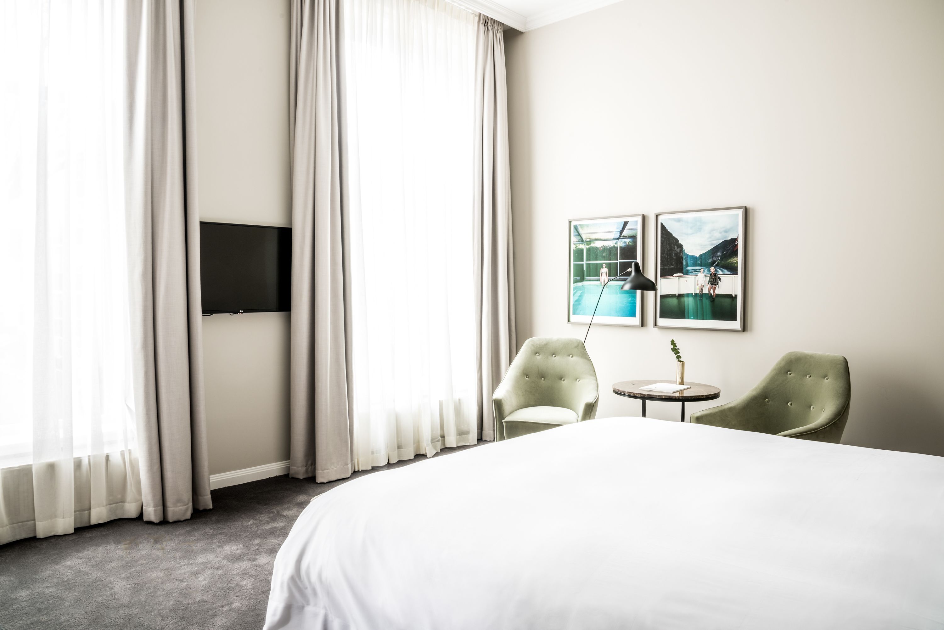A hotel room with a large bed, two armchairs, a small table, a flat-screen TV mounted on the wall, and two framed pictures depicting outdoor scenes.