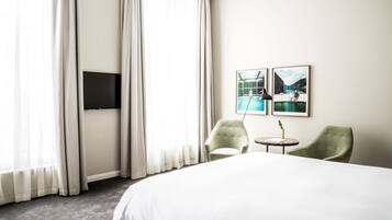 A hotel room with a large bed, two armchairs, a small table, a flat-screen TV mounted on the wall, and two framed pictures depicting outdoor scenes.