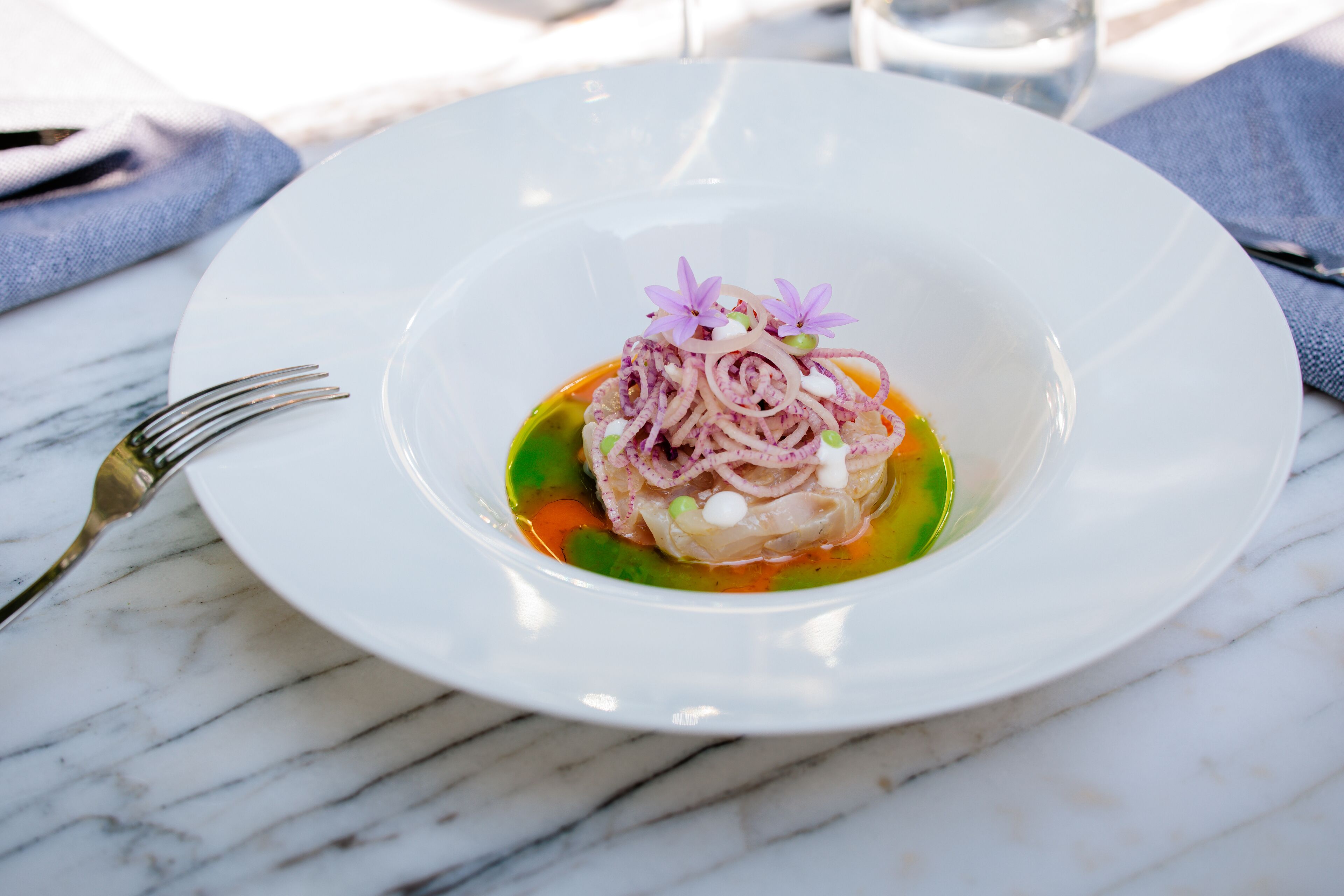 Un plato blanco con un plato de mariscos, un tenedor y un vaso de agua sobre una mesa de mármol.