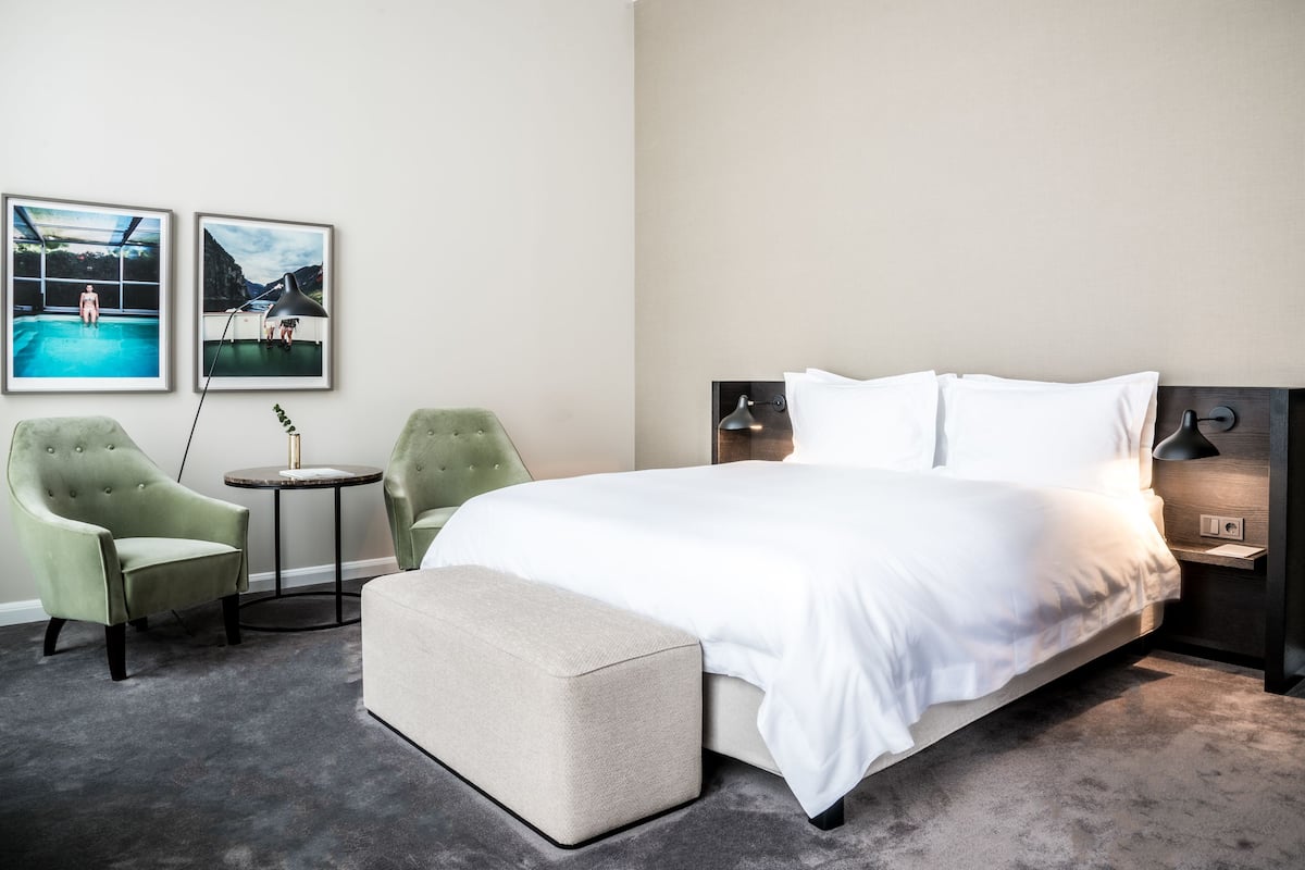 Une chambre d’hôtel avec un grand lit, deux fauteuils, une petite table ronde et deux tableaux encadrés au mur.