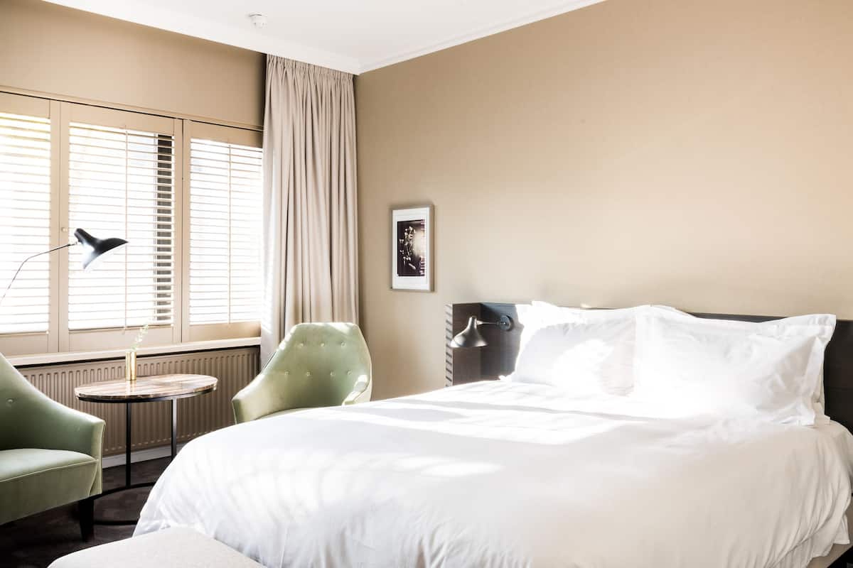 Une chambre d’hôtel avec un grand lit, une table ronde, un fauteuil vert et une fenêtre avec des stores.