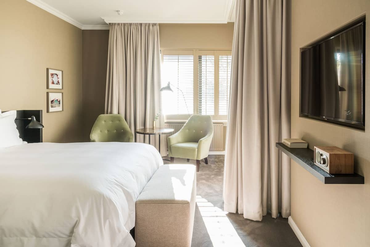 Une chambre d’hôtel avec un grand lit, deux chaises, une télévision et une fenêtre avec des rideaux.
