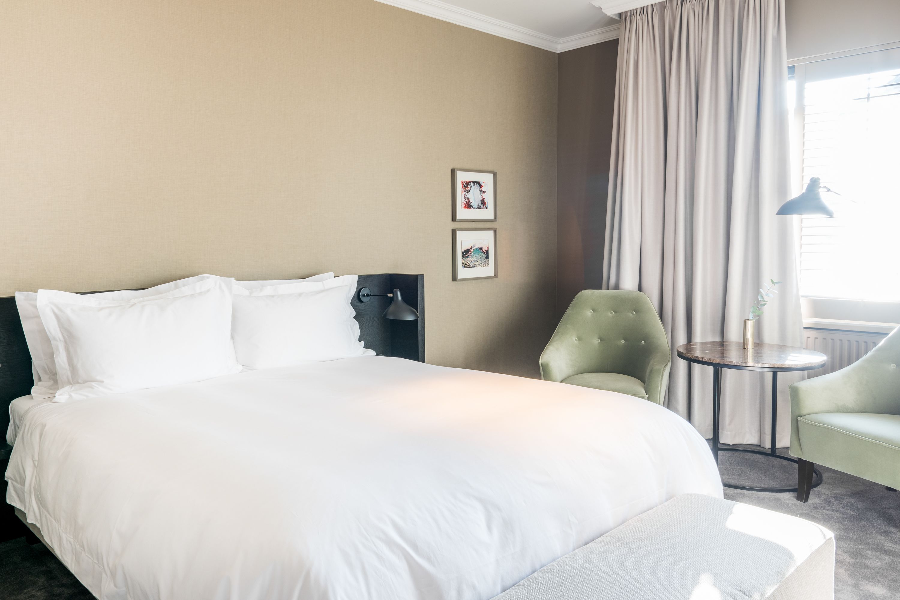 A hotel room with a large bed, two framed pictures on the wall, a green armchair, a small round table, and a window with curtains.