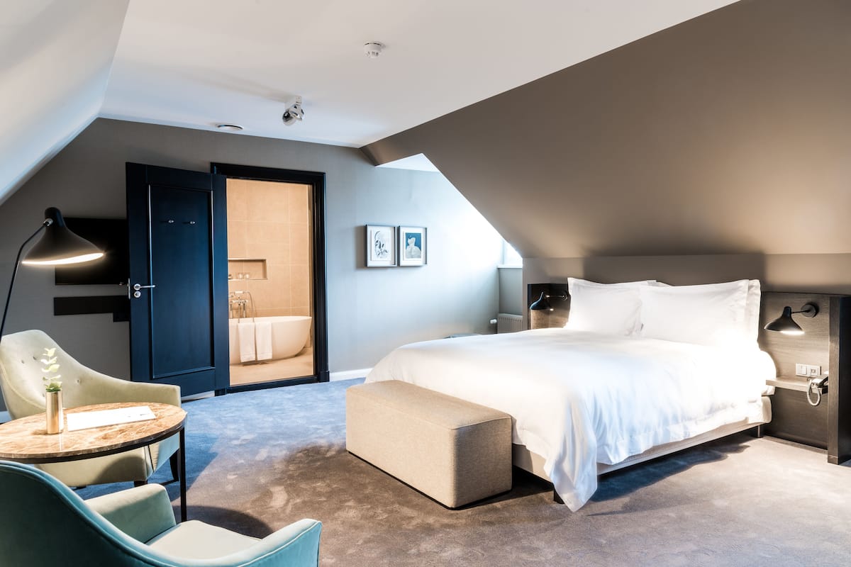Une chambre moderne avec un grand lit, un bureau et une chaise. On y trouve une porte donnant sur une salle de bain, ainsi qu’une petite table avec une lampe.