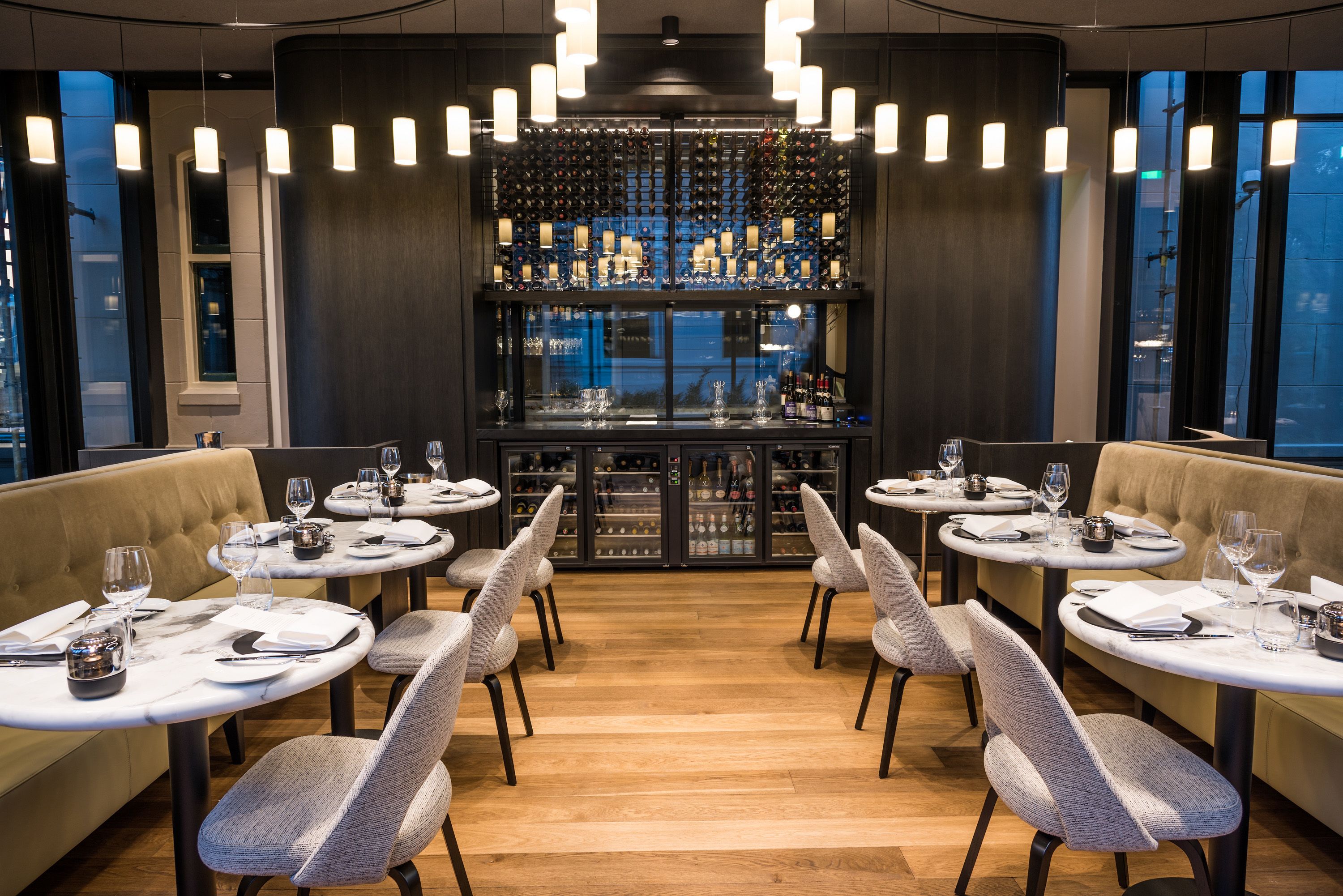 A restaurant with round tables, grey upholstered booths, and a wine rack in the background.