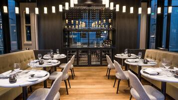 A restaurant with round tables, grey upholstered booths, and a wine rack in the background.