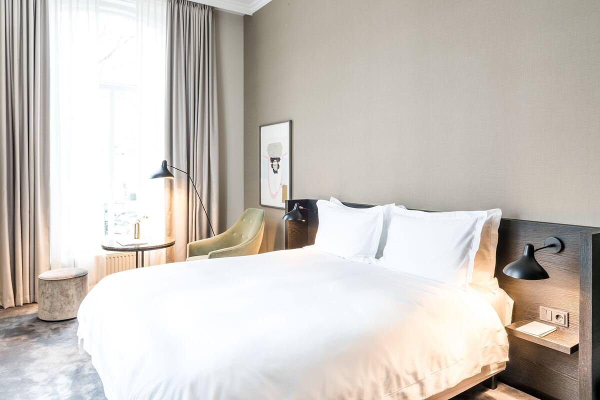 Une chambre d’hôtel avec un grand lit, une table de chevet, une chaise, une lampe et une fenêtre avec des rideaux.