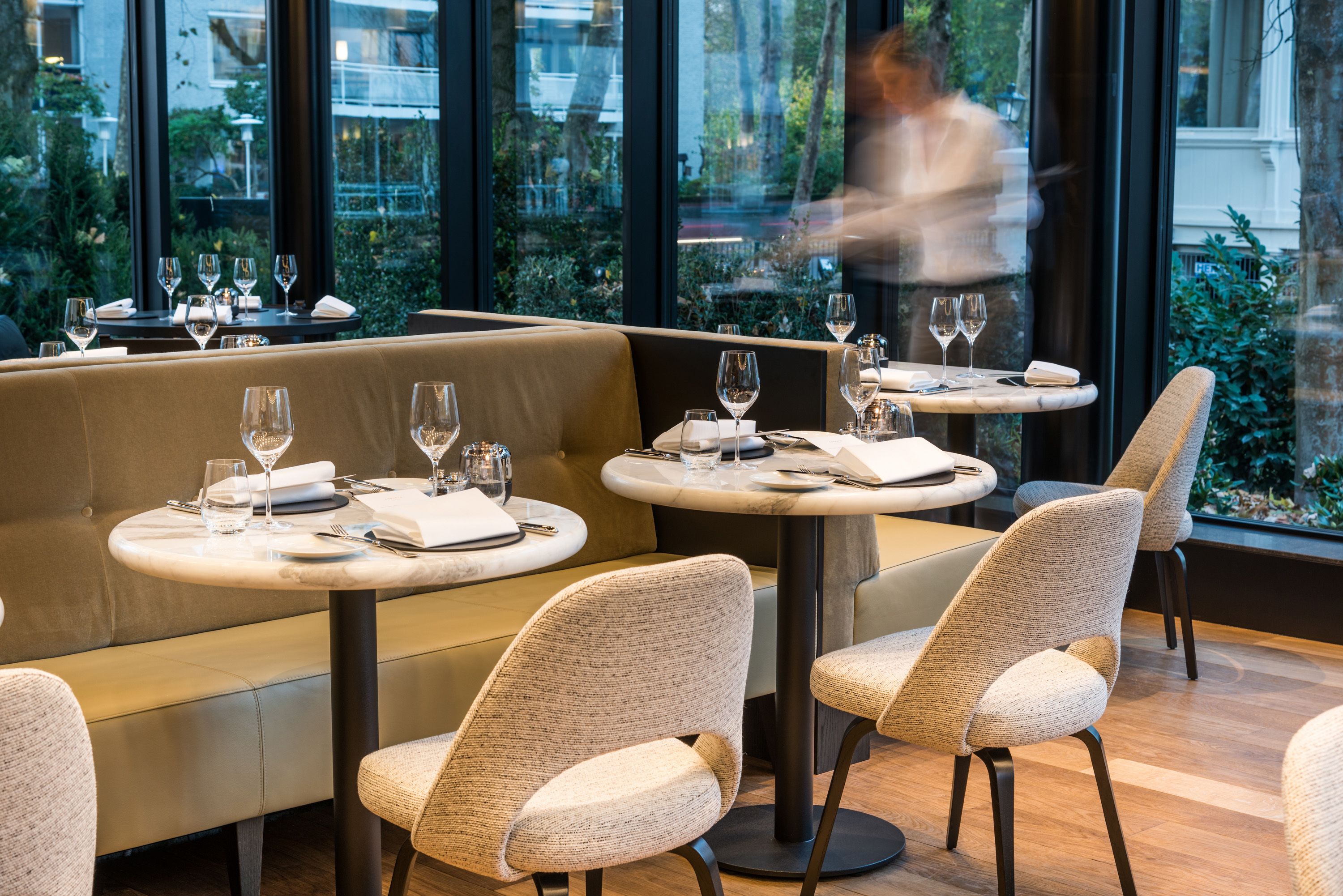 Un restaurant avec des tables rondes et des sièges rembourrés, de grandes fenêtres donnant sur un paysage verdoyant.