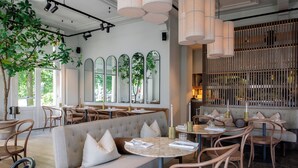 A restaurant with a modern interior, featuring a mix of seating options including a central round table, a sofa with cushions, and individual chairs. There are large windows allowing natural light to enter, and the space is decorated with plants and mirrors. - Pillows Luxury Boutique Hotel Aan De IJssel (Deventer)