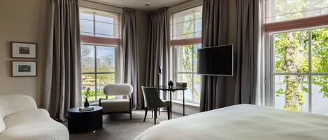 A modern hotel room with a large bed, a seating area with a chair and a small table, a flat-screen TV mounted on the wall, and large windows offering a view of the outdoors.
