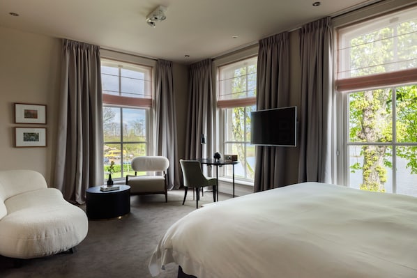 Una habitación de hotel moderna con una cama grande, un área de descanso con una silla y una mesita, un televisor de pantalla plana en la pared y ventanas amplias con vistas al exterior.
