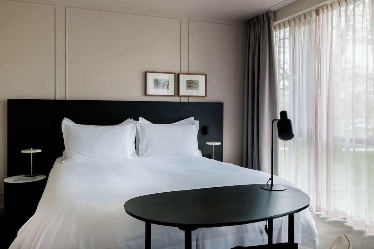 Habitación de hotel con una cama grande, dos mesitas de noche, una mesa redonda y vistas a árboles a través de cortinas translúcidas.
