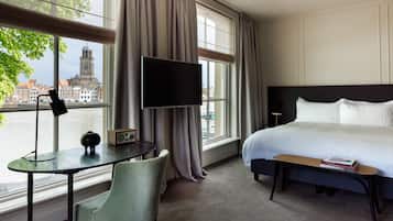 A hotel room with a bed, a desk with a chair, a TV, and a view of a cityscape with a tower.