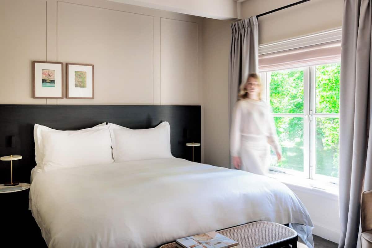 Una habitación de hotel con una cama grande, una mesita de noche con lámpara y una ventana con cortinas.