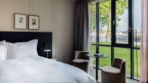 A hotel room with a large bed, two framed pictures on the wall, a small round table, two chairs, and a view of a grassy area and buildings through large windows. - Pillows Luxury Boutique Hotel Aan De IJssel (Deventer)