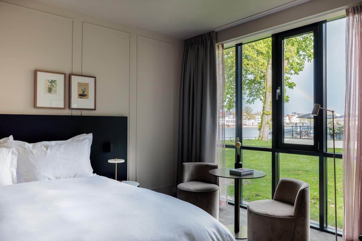 Habitación de hotel con una cama grande, dos cuadros en la pared, una mesita redonda, dos sillas y vistas a un área verde y edificios a través de amplios ventanales.