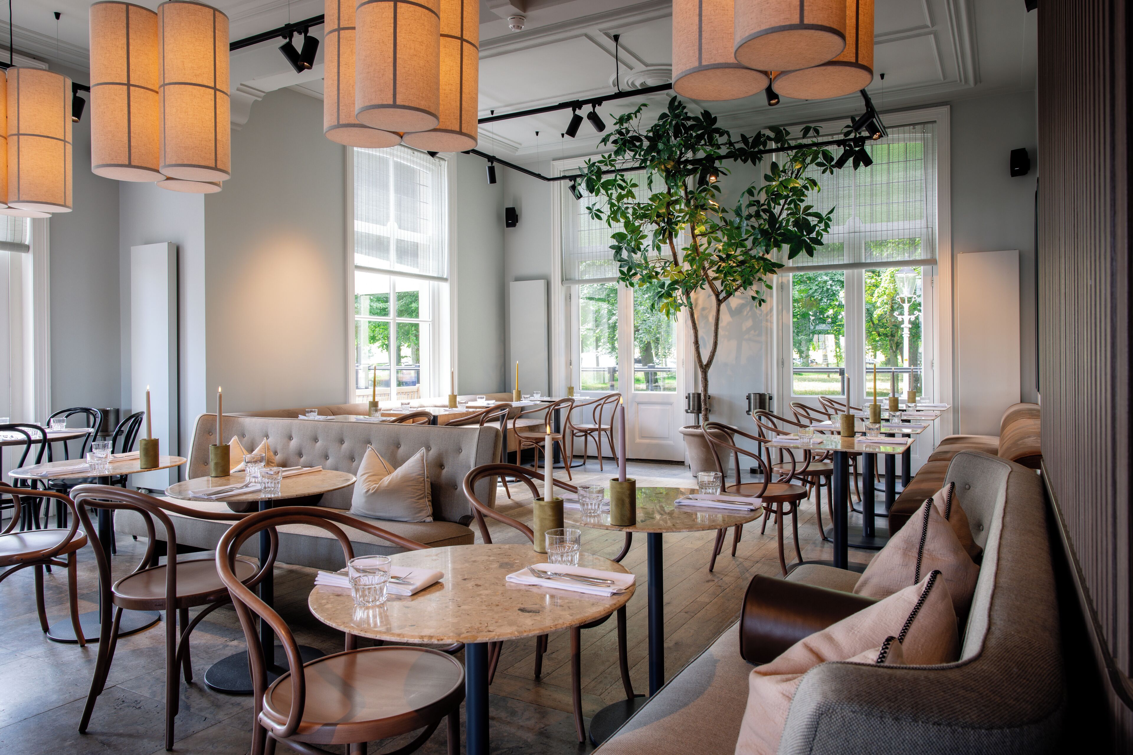 A modern restaurant interior with round tables, cushioned seating, pendant lighting, and large windows.