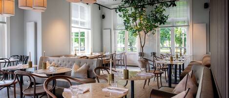 Un interior de restaurante moderno con mesas redondas, asientos acolchados, iluminación colgante y amplios ventanales.