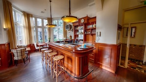 A bar area with wooden bar stools, a wooden counter, and hanging pendant lights. - Fletcher Hotel Gilde (Deventer)