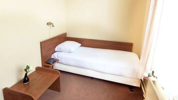 Een kleine kamer met een enkel bed, een houten hoofdeinde, een nachtkastje met een vaas en een radiator.