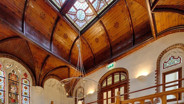 A grand wooden staircase with intricate designs, a high ceiling with stained glass windows, and a second-floor balcony. - Fletcher Hotel Gilde (Deventer)