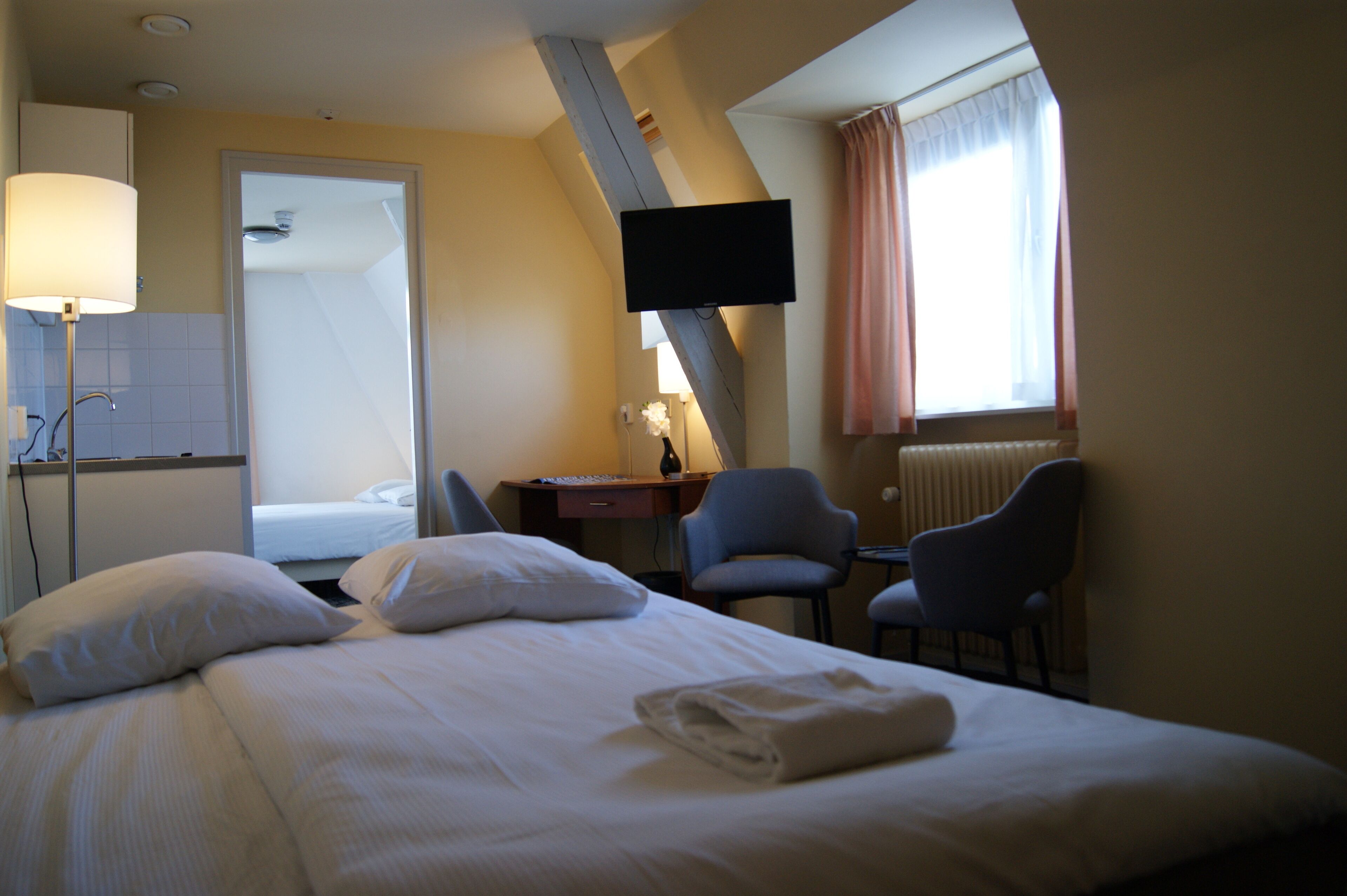 Ett hotellrum med en säng, ett skrivbord, två stolar, en TV och ett fönster med gardiner.