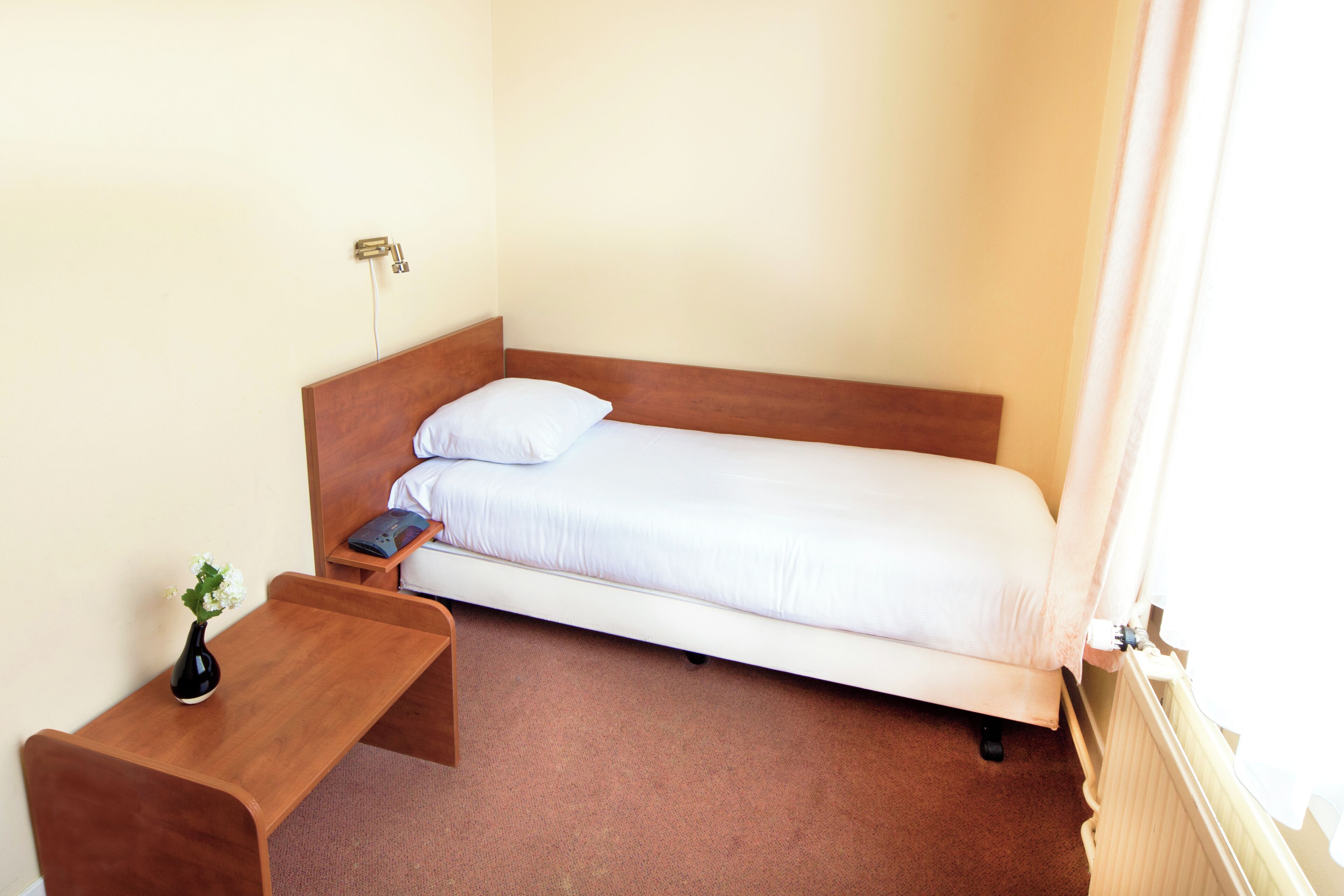 A small, single-bed room with a wooden headboard, a bedside table with a vase, and a radiator.