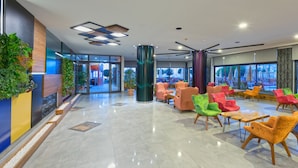 A modern lobby with colorful chairs, a central pillar, and a view of the outside. - Monart City Hotel - All Inclusive (Alanya)