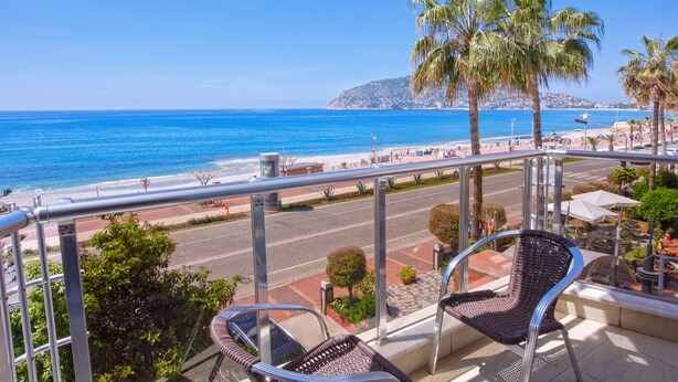 A balcony with wicker chairs overlooking a beach with palm trees and a clear blue sky. - Monart City Hotel - All Inclusive (Alanya)