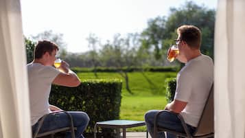 Dos personas sentadas en una pequeña mesa al aire libre, con un seto y follaje de fondo.