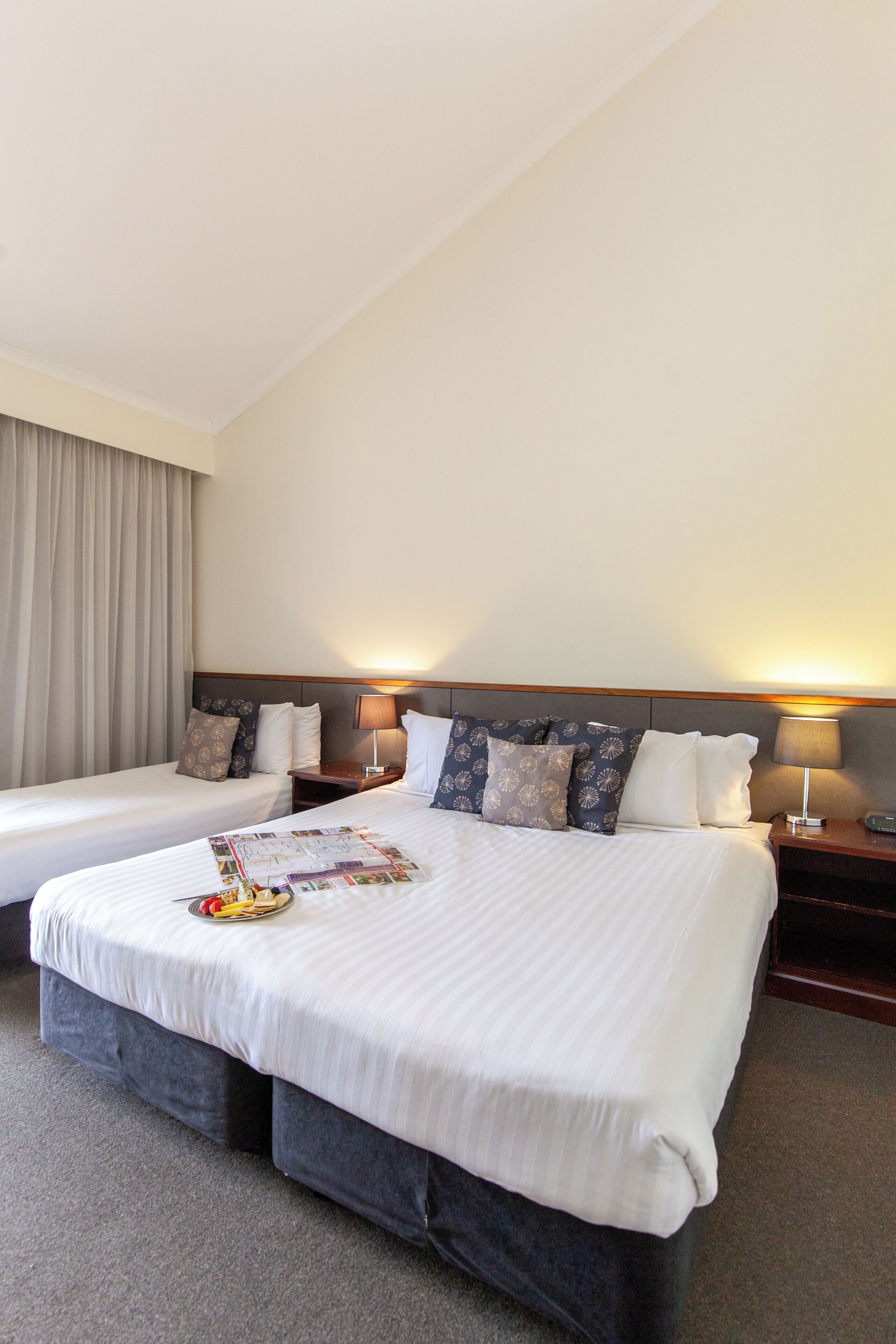 A hotel room with two beds, a nightstand, a lamp, and a window with curtains.