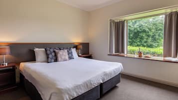 A hotel room with a large bed, two bedside tables, a window with curtains, and a view of greenery outside.