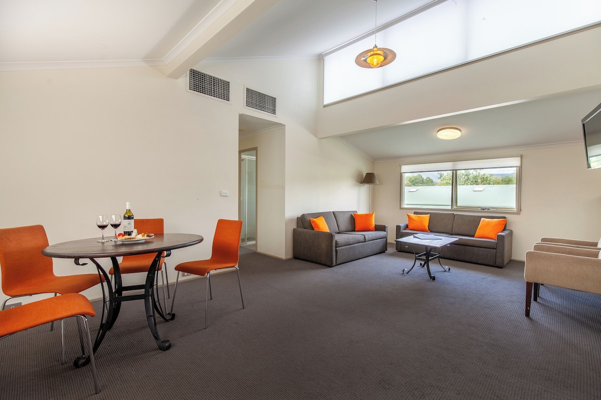 A modern lounge with a round table, orange chairs, a grey sofa with orange cushions, and a large window providing natural light.