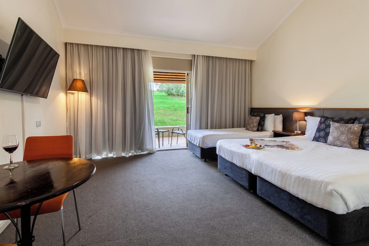 A hotel room with a bed, a TV, a table with a glass of wine, and a view of the outdoors.