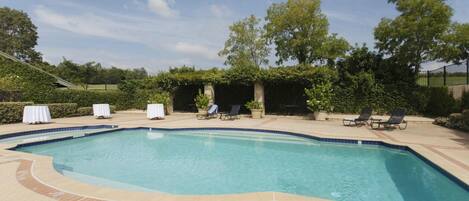 A rectangular swimming pool with a tiled deck, surrounded by lounge chairs and tables, and a pergola with climbing plants.
