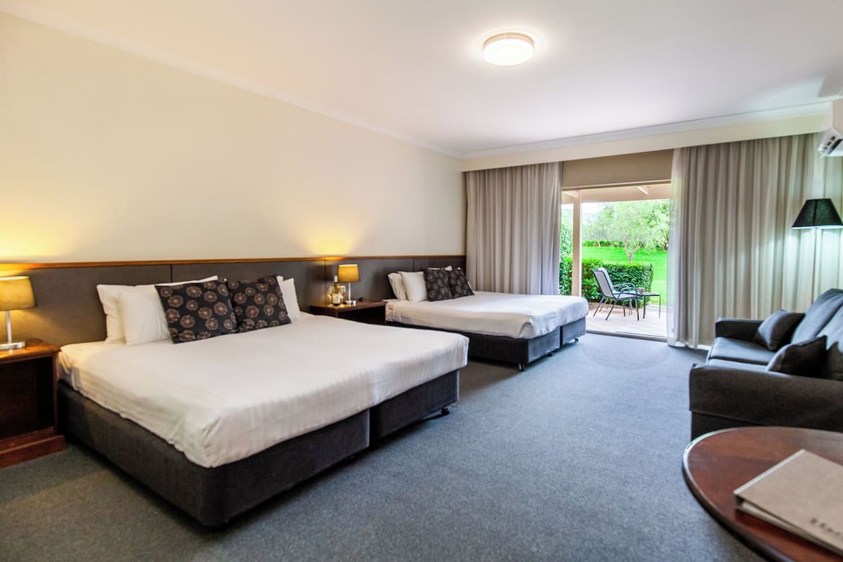 A hotel room with two beds, a sofa, a table, and a view of a garden.