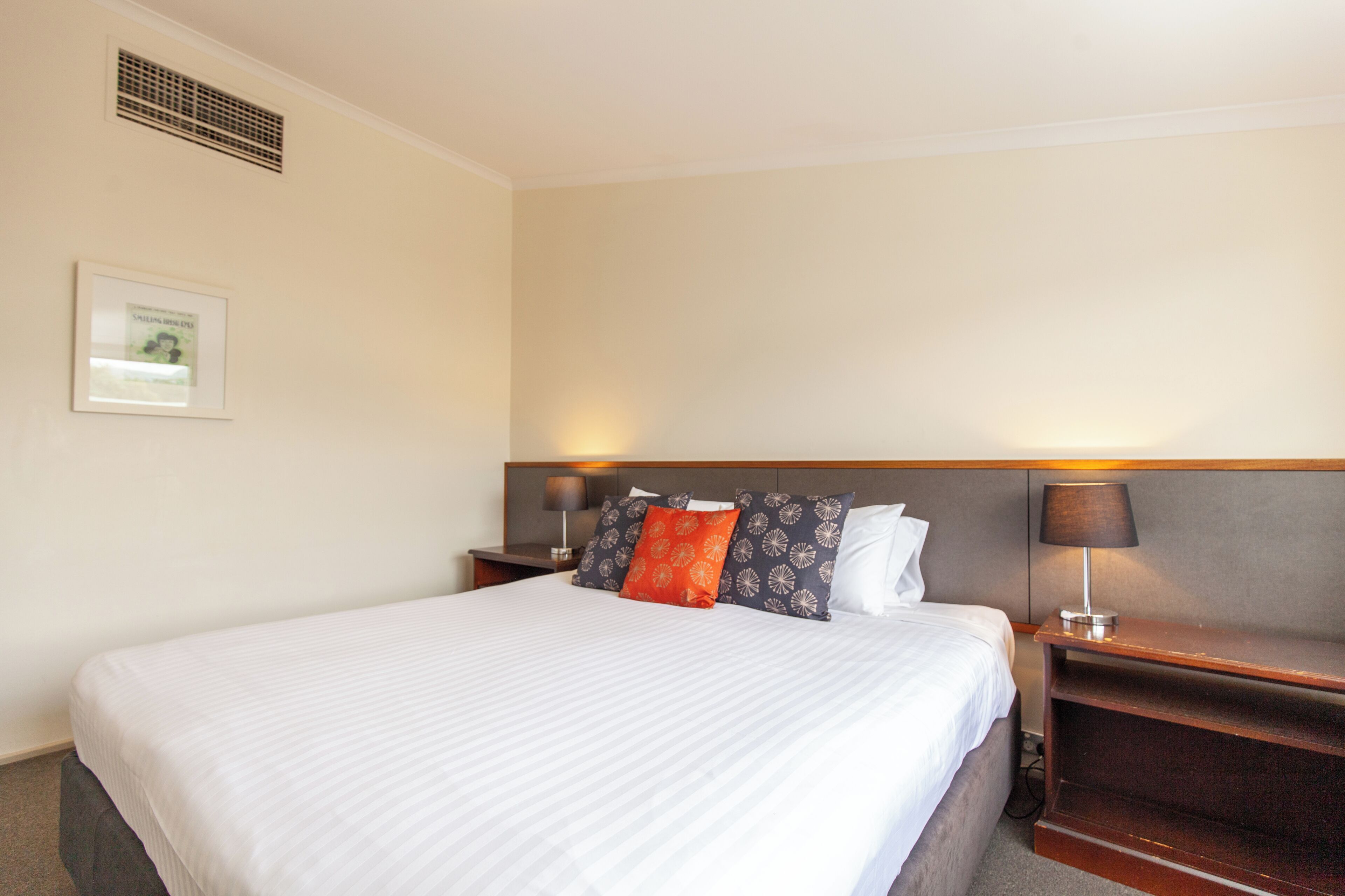 A hotel room with a bed, two bedside tables, a nightstand, a wall-mounted lamp, and a framed picture on the wall.