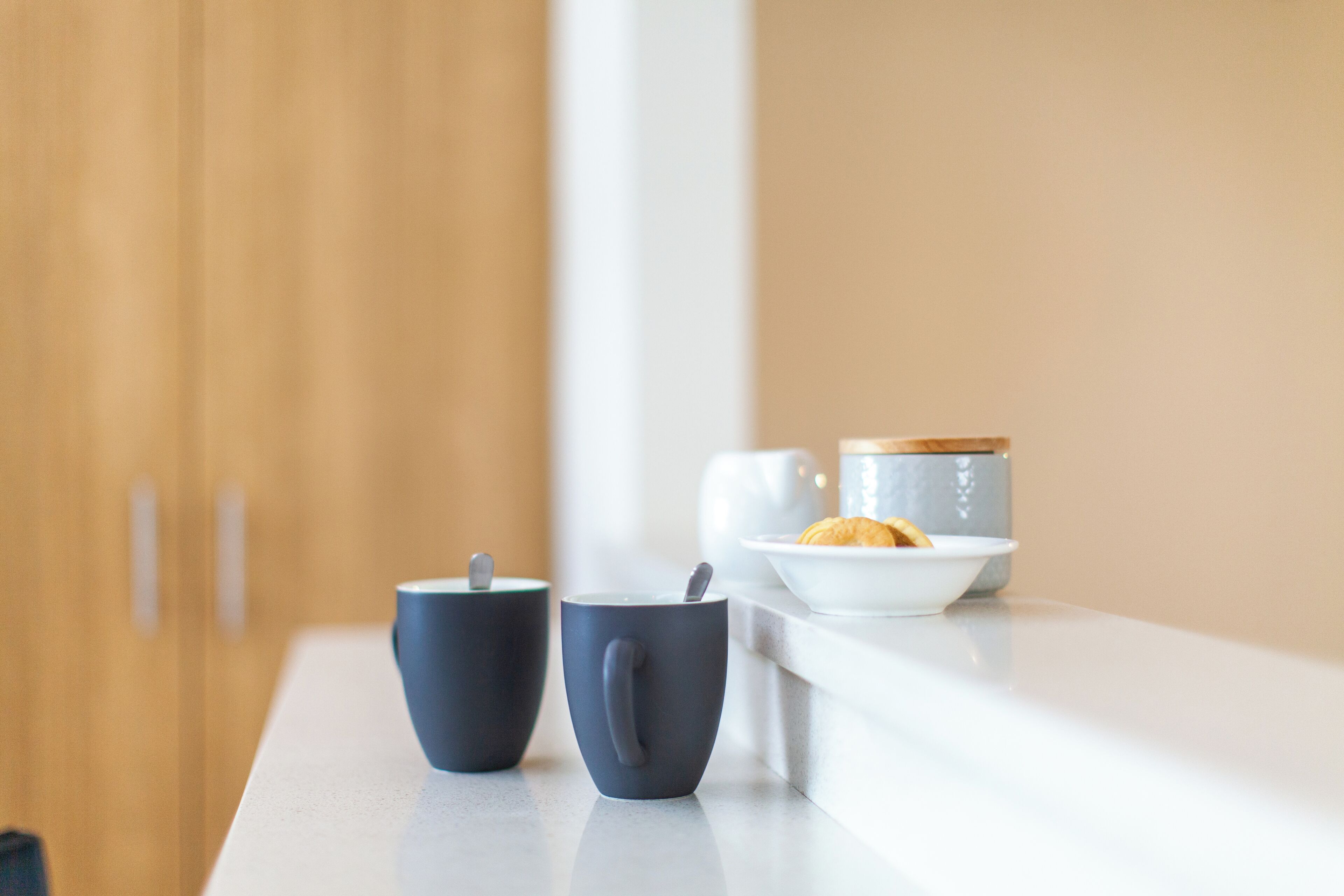 A modern kitchen with a white countertop, two dark mugs, a white teapot, and a bowl with a pastry.