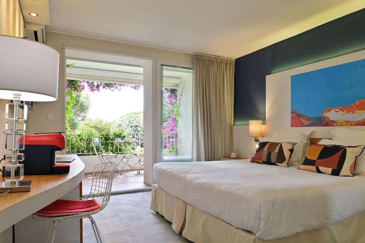 Habitación de hotel con cama, escritorio y silla. Tiene una ventana con vistas a zonas verdes y un balcón.