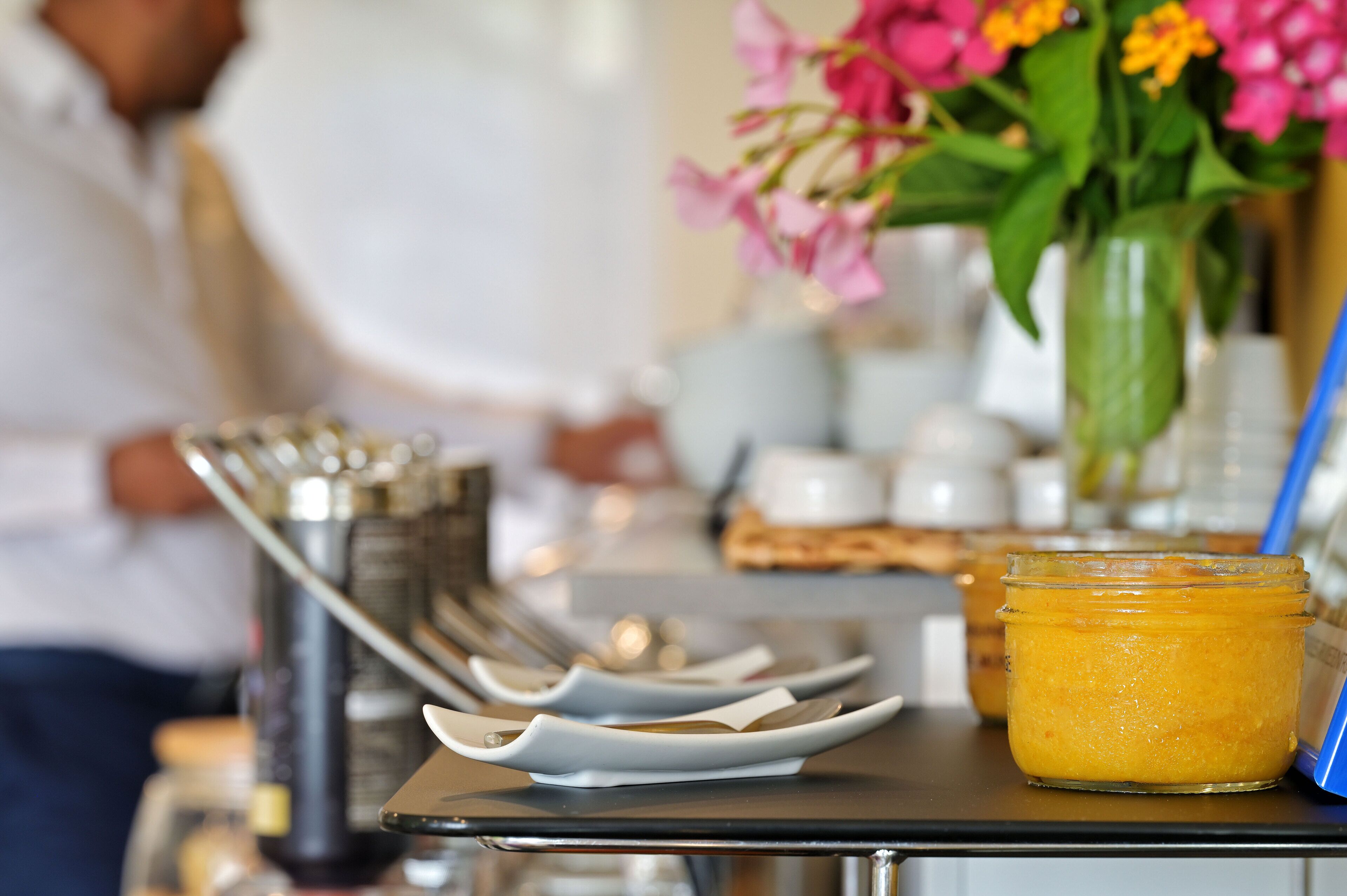 A table with a jar of yellow jam, a vase with pink flowers, and a blue napkin holder.