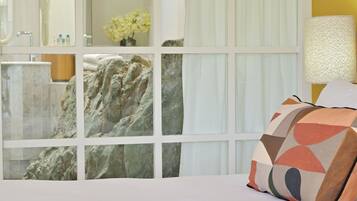 Una habitación de hotel con cama, un ventanal con vista a un baño y una formación rocosa, y una lámpara en la mesita de noche.