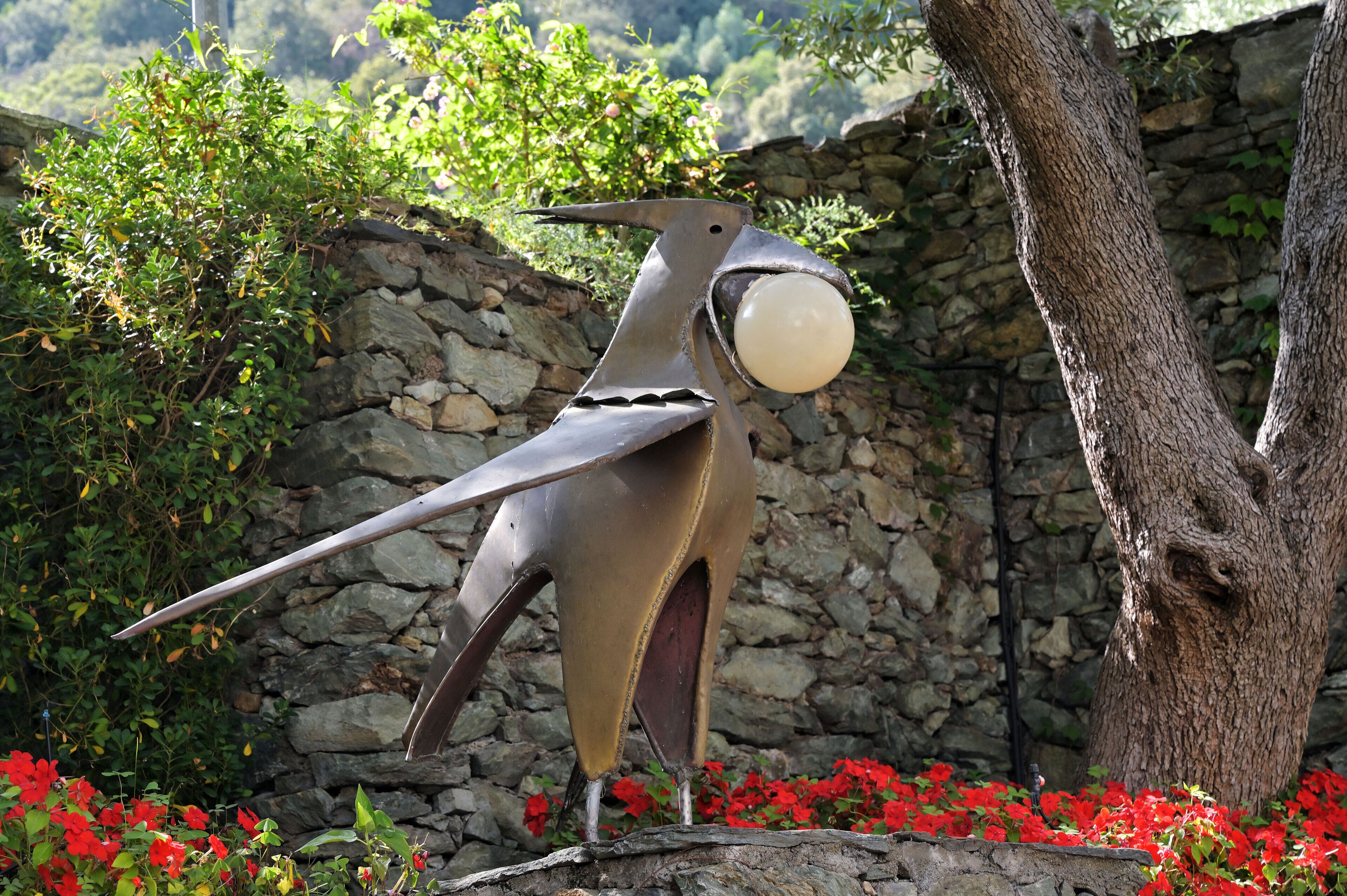 Escultura d'un ocell amb un ou blanc al bec, davant d'una paret de pedra i envoltat de flors vermelles.