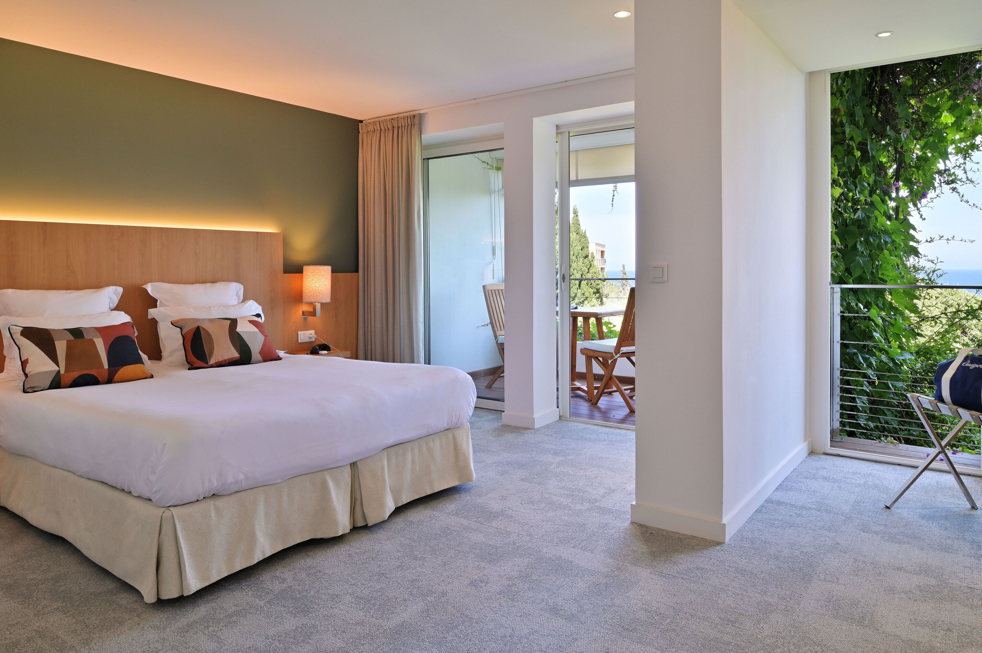 Habitación de hotel con una cama grande, una mesita de noche, una lámpara y vistas a un balcón con sillas.