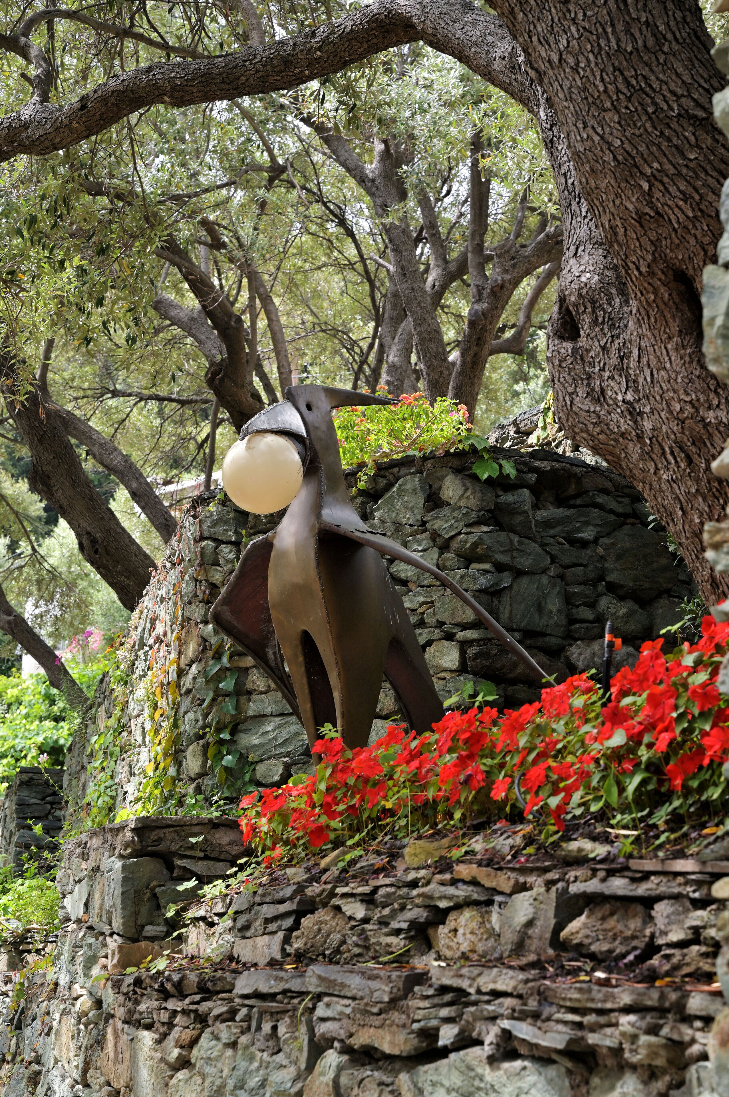 A sculpture of a bird with a glowing orb, set against a stone wall with red flowers.