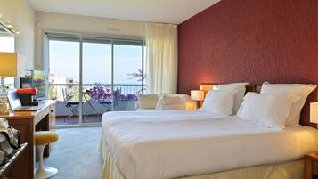 A hotel room with a large bed, a desk with a laptop, a chair, and a view of the sea through a balcony.